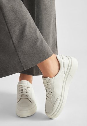 Bílé kožené tenisky s tlustou podrážkou nošené s šedými širokými kalhotami na světlém pozadí.