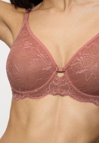 Reggiseno in pizzo rosa con scollo a V, caratterizzato da motivi floreali, spalline regolabili e un dettaglio a fiocco centrale. Banda elastica liscia nella parte inferiore.