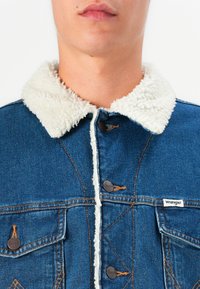 Denim jakna u plavoj boji s bijelim krznom na ovratniku. Sadrži smeđu šavovinu i metalne gumbe. Ima dva prednja džepa. Vidljiva oznaka marke Wrangler.
