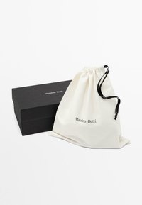 Boîte noire Massimo Dutti à côté d’un sac blanc à cordon noir, tous deux avec le logo Massimo Dutti sur un fond blanc uni.