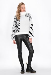 Pulover pufoș alb cu imprimeuri de leopard și zebra negre, guler înalt, mâneci balon; purtat cu pantaloni din piele neagră lucioasă și cizme cu șireturi.