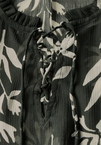 Grüne Bluse mit Blumenmuster, die einen Schnürrëntelkragen, strukturierte Stoffe und einen rüschigen Kragen mit weißen Blättern auf dunklem Hintergrund zeigt.
