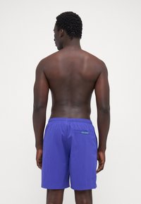 Purple swim shorts made of lightweight fabric. Features an elastic waistband and a small back pocket with green branding.