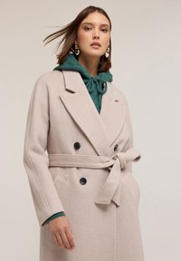 Abrigo rosa claro de doble botonadura, hecho de un tejido suave con cintura ceñida. Presenta botones negros y un suéter con capucha verde debajo.