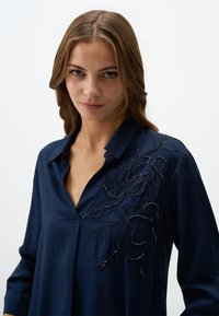 Blusa blu navy con scollatura a V e maniche lunghe, caratterizzata da ricami floreali neri con dettagli di perline sul lato sinistro. Tessuto morbido.