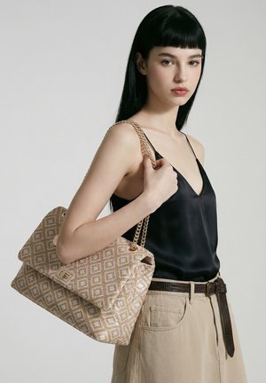 Jeune femme aux longs cheveux foncés porte un caraco en satin noir et un pantalon beige, tenant un sac à main beige à motif avec une chaîne dorée sur l'épaule.
