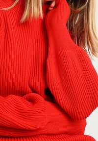 Roter gerippter Pullover mit langen, voluminösen Ärmeln und einem dicken, strukturierten Stoff. Die Ärmelbündchen sitzen eng am Handgelenk.