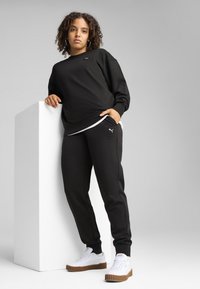 Svart sweatshirt och byxset i mjukt material, med en liten logotyp, i kombination med vita sneakers med texturerad sula, modell som lutar sig mot en vit pelare.