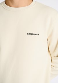 Sudadera color crema hecha de una tela suave, con mangas largas y cuello redondo. "LINDHBERGH" está bordado en negro en el pecho.