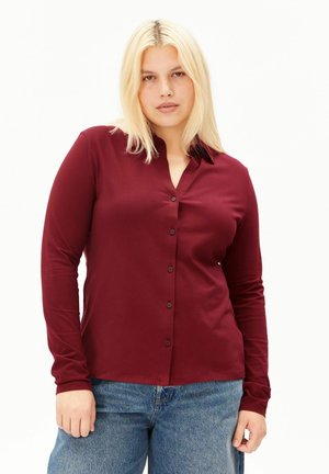 Frau mit schulterlangen blonden Haaren, die ein geknöpftes langärmeliges bordeauxrotes Hemd und blaue Jeans vor weißem Hintergrund trägt.