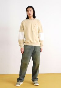 Sweatshirt beige con maniche bianche, tasca sul petto e polsini a coste. Indossato con jeans larghi verde scuro e sneakers marroni.