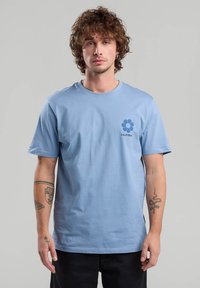 Vaaleansininen puuvillainen t-paita, jossa on lyhyet hihat, pyöreä kaula-aukko ja etupuolella kukkakuva grafiikka, jossa lukee "CALIFORNIA". Yksinkertainen, rento malli.