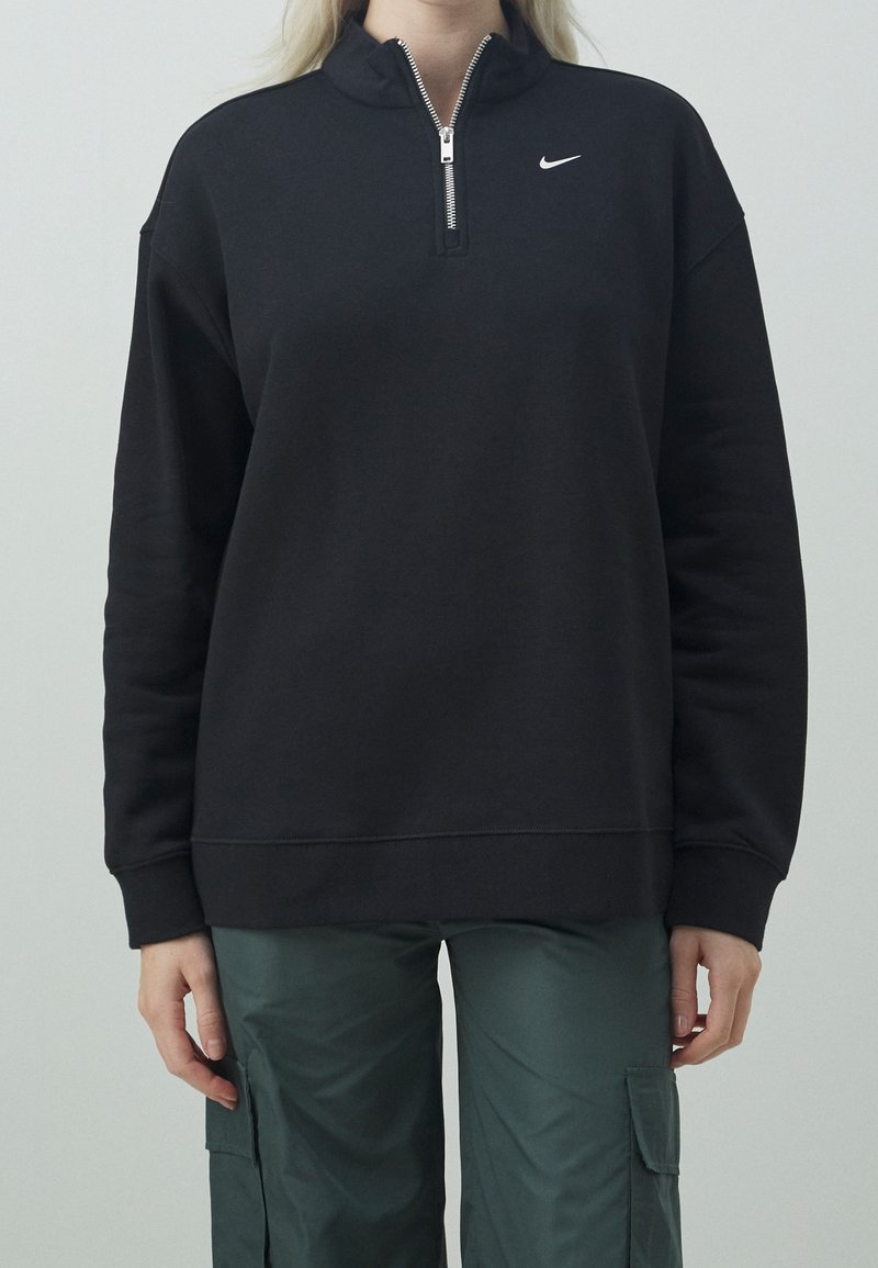 Pull à col montant noir en polaire, avec une demi-fermeture éclair et un petit logo blanc sur la poitrine. Le tissu est lisse avec une coupe décontractée.