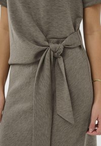 Sivina obleka s kravato spredaj, izdelana iz mehke, teksturirane tkanine, z detajlom z vozlom v pasu. Vključuje ustrezne kravate za nastavljiv fit.