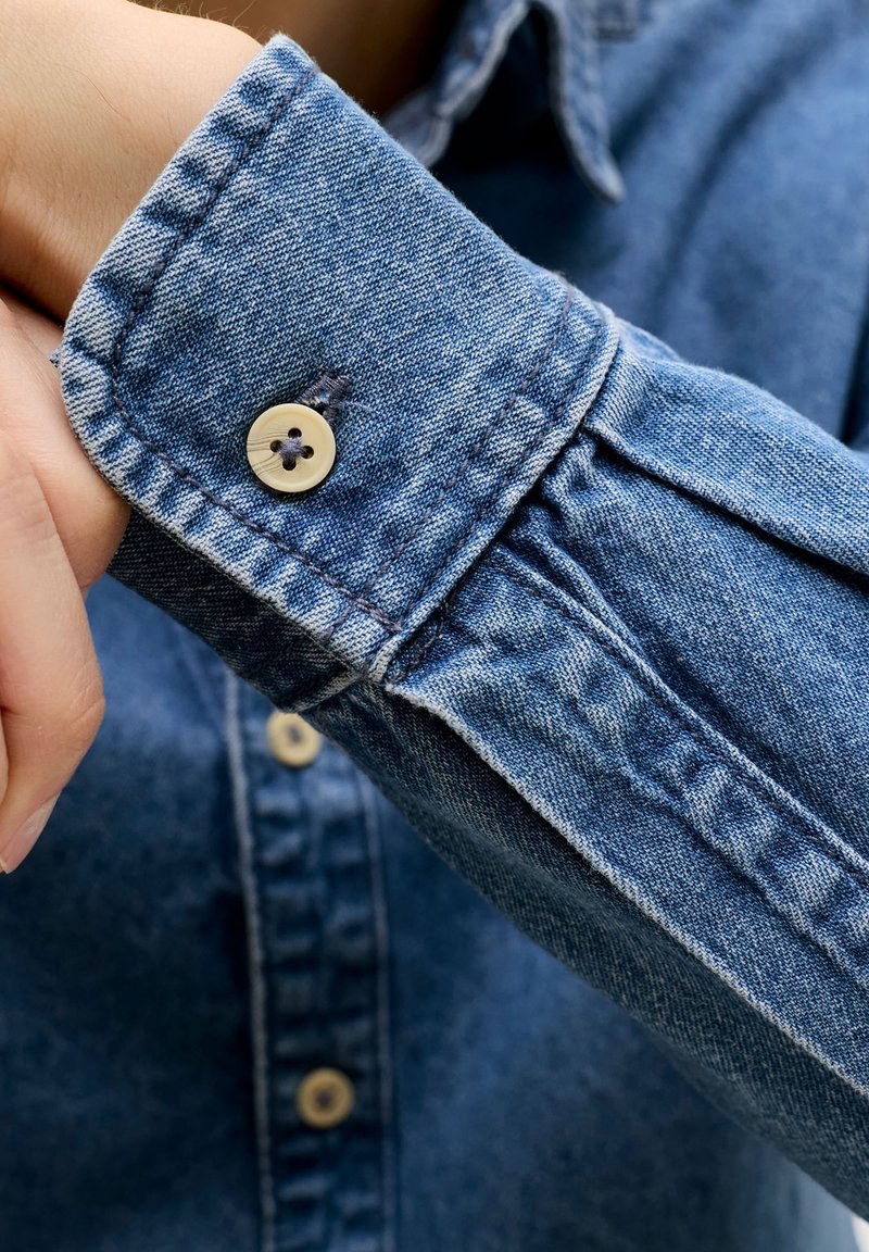 Άτομο κουμπώνει το μανίκι μπλε τζιν πουκάμισου, εμφανίζοντας κοντινό πλάνο του χεριού και λεπτομερή ραφή με μπεζ κουμπιά.