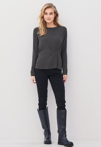 Blonde woman standing against white wall wearing dark gray long-sleeve blouse, black fitted pants, and knee-high black boots.