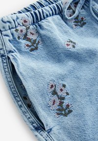 Lichtblauwe denim stof met geborduurde witte en rode bloemen, met een elastische tailleband en een opening voor een zijzak.