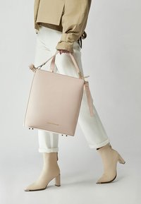 Neliönmuotoinen, vaaleanpunainen nahkalaukku, jossa on lyhyet kahvat, säädettävä olkahihna, kultaiset metalliosat ja hillitty logo. Yhdistetty valkoisiin housuihin ja beigen värisiin korollisiin saappaisiin.