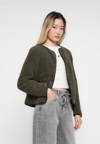 Olivengrønn, teksturert fleecejakke med rund hals, svart kant og trykknapper, kombinert med en hvit topp og grå løstsittende jeans.
