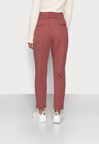 Person som bär rostfärgade tapered-byxor med elastisk midja och vita Adidas-sneakers, stående mot en neutral bakgrund.