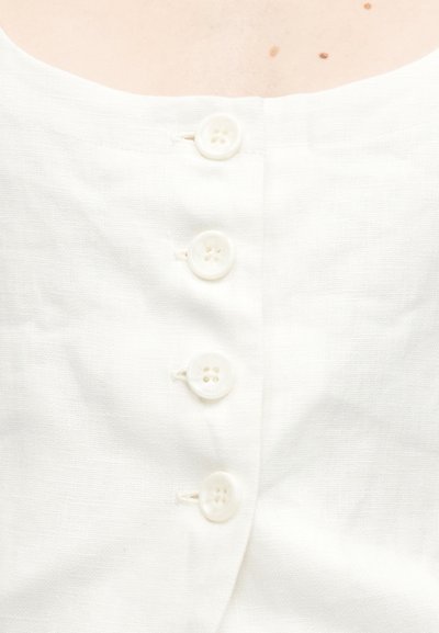 Λευκό λινό ένδυμα με στρογγυλή λαιμόκοψη, διακοσμημένο με πέντε στρογγυλά κουμπιά με τέσσερις τρύπες το καθένα κατά μήκος του εμπρός. Η υφή του υφάσματος είναι ελαφρώς τσαλακωμένη.