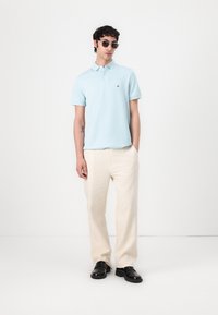 Camisa polo de color azul claro con mangas cortas, que presenta un pequeño logo. Pantalones sueltos de color crema y zapatos negros completan el conjunto.