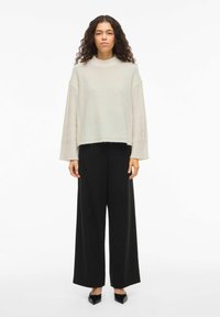 Helebeež kudumiga kõrge kaelusega kampsun, millel on laiade varrukatega ja töödeldud äär, sobitatud kõrge vöökohaga mustade laiade pükstega.