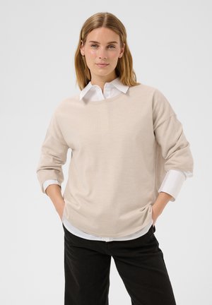 Femme aux cheveux raides châtain clair porte un pull beige sur une chemise blanche à col et un pantalon noir, debout les mains dans les poches.