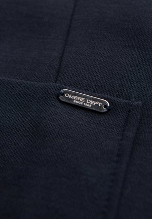 Tmavě modrá pletená látka s malou obdélníkovou kovovou štítkem s rytým nápisem "OMBRE DEPT Since 1989" přišitou na ní.