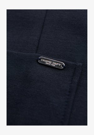 Tela de punto azul oscuro con una pequeña etiqueta metálica rectangular grabada con "OMBRE DEPT Since 1989" cosida sobre ella.