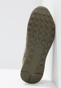 Semelle d'une basket Nike vert olive avec des motifs carrés et un logo visible sur la semelle extérieure, photographiée en gros plan sur fond blanc.