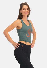 Grünes Baumwoll-Tanktop mit einem geknoteten Saum und orangefarbigem "Om"-Symbol; kombiniert mit hoch taillierten schwarzen Leggings mit einem goldenen Akzent-Logo.