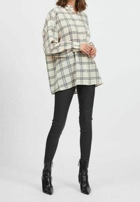 Woman wearing oversized cream plaid shirt, black fitted pants, and black heeled ankle boots, standing with hands clasped in front.