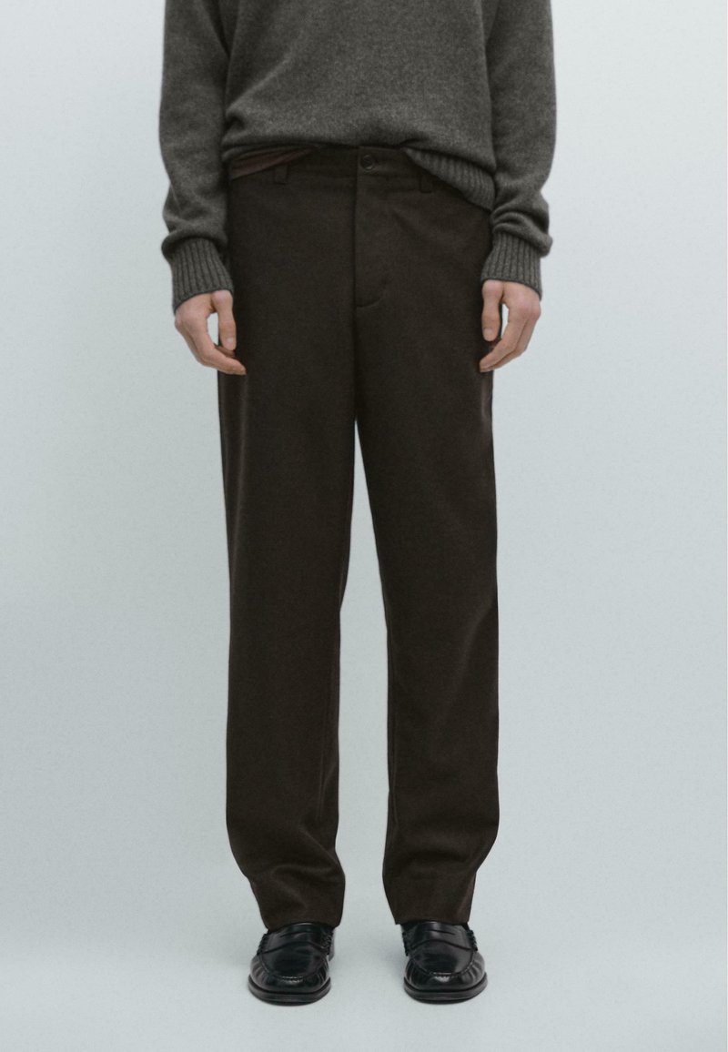 Brązowe wełniane spodnie o prostym kroju, z przednimi kieszeniami i subtelną fakturą, w połączeniu z czarnymi eleganckimi butami i szarym swetrem.