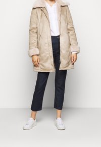 Abrigo beige de piel sintética con cuello y puños suaves, cierre de cremallera, llevado sobre una camisa blanca y jeans oscuros recortados, combinado con zapatillas blancas.