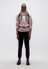 Γυναίκα που φοράει γυαλιά ηλίου, ένα ροζ μπουφάν Columbia, μαύρο παντελόνι, παπούτσια πεζοπορίας και ένα ανοιχτό πράσινο σαμαράκι, πάνω σε απλό λευκό φόντο.