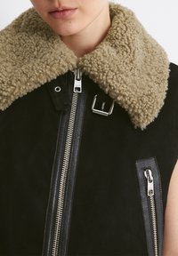 Melnas šerfē vestes ar lielu beigu krāsas apkakli, metāla rāvējslēdzēju un ādas akcentiem, struktūrēta dizaina un mīksta tekstūra.