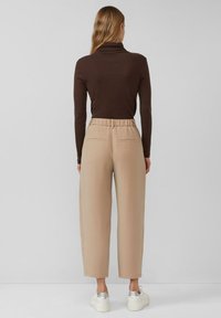 Braunes Langarmoberteil mit Stehkragen, kombiniert mit beigen, weit geschnittenen Hosen mit Kordelzug an der Taille und Gesäßtaschen. Weiße Schuhe mit silbernen Akzenten.