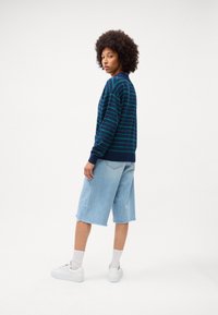 Temno modra pulover s črtami v zeleni barvi, v kombinaciji s svetlo modrimi denim bermuda kratkimi hlačami. Model nosi bele športne copate in kratke bele nogavice.
