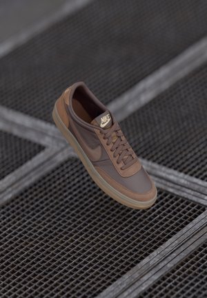Brown Nike sneaker with laces and logo, placed on a metal grid floor with diagonal lines, shown at an angled top view.