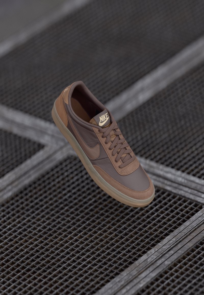 Brown Nike sneaker with laces and logo, placed on a metal grid floor with diagonal lines, shown at an angled top view.