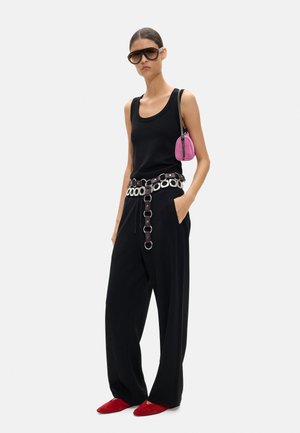 Kobieta ubrana w czarny bezrękawnik, szerokie czarne spodnie z paskiem z srebrnymi ogniwami, czerwone buty, oversize'owe okulary przeciwsłoneczne i małą różową torbę na ramię.