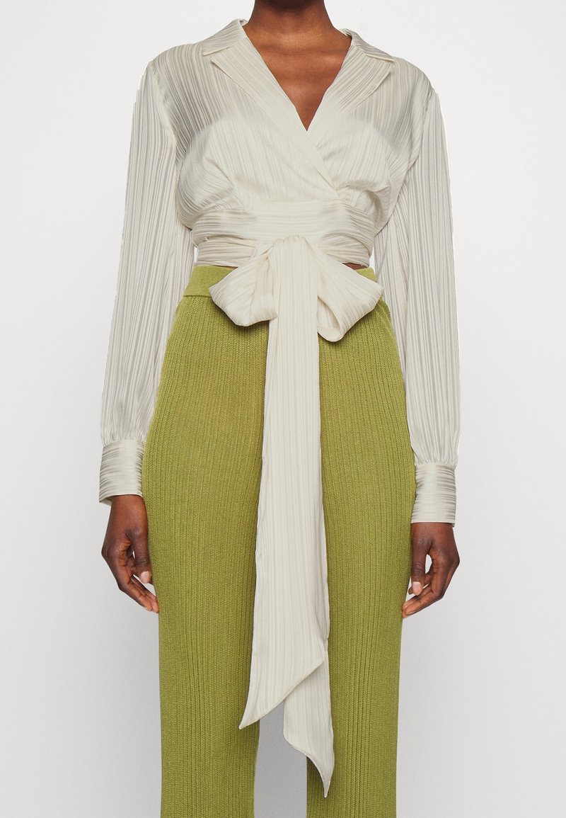 Blouse croisée ivoire à manches longues avec plis verticaux, détail noué à la taille et col structuré, associée à un pantalon taille haute texturé vert olive.