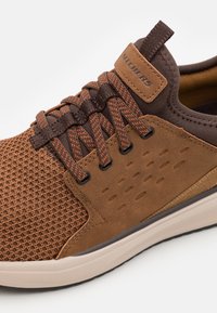 Basket de sport marron avec une tige en mesh texturé, un col rembourré, des lacets contrastants et une semelle en caoutchouc avec des accents subtils et une ventilation.