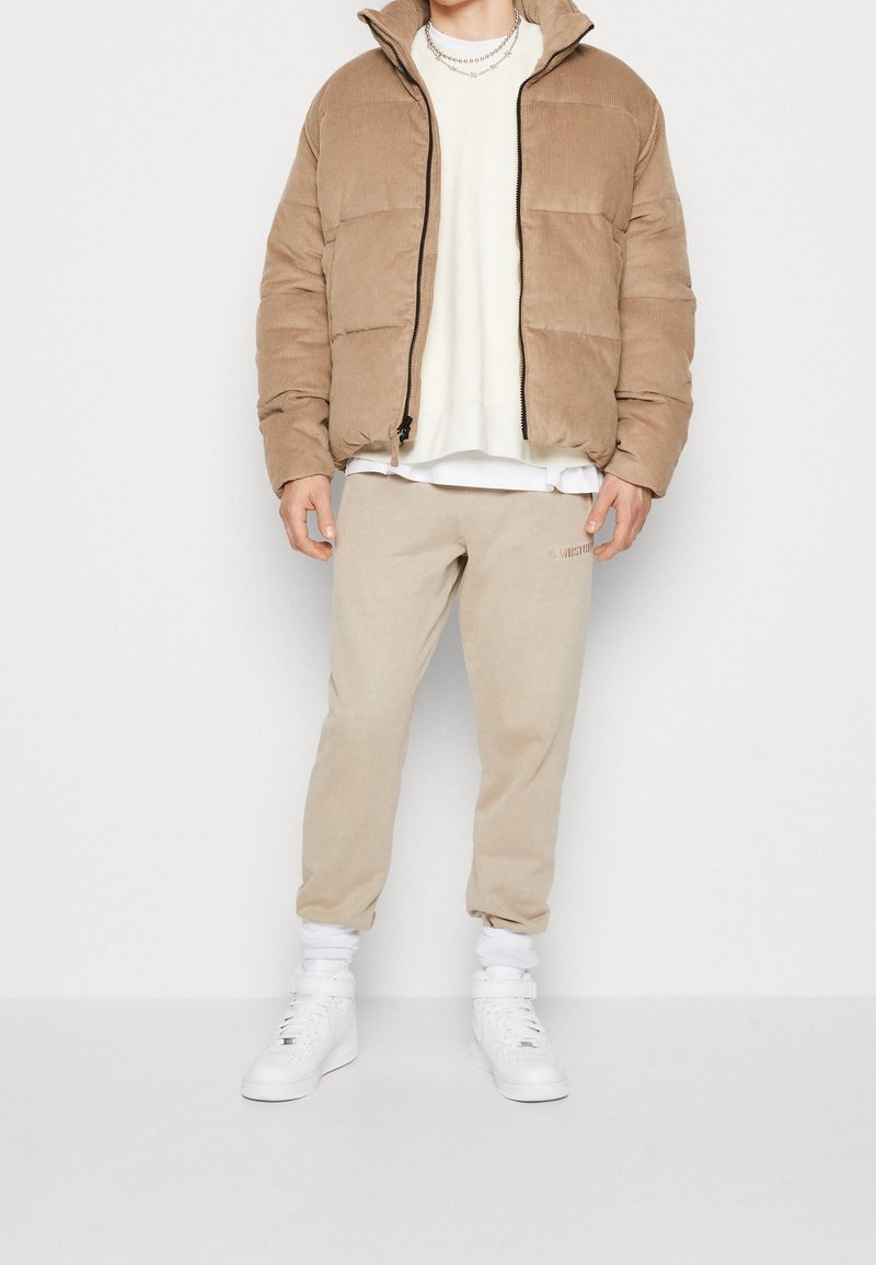 Personne portant une veste matelassée beige, un sweat crème, un pantalon de survêtement beige, des chaussettes blanches et des baskets blanches montantes, debout devant un fond uni.