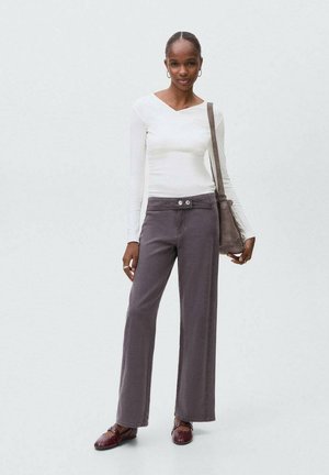 Vrouw staat in een witte lange mouw top, grijze wijde broek met knopen, bordeauxrode schoenen en draagt een bruine schoudertas.