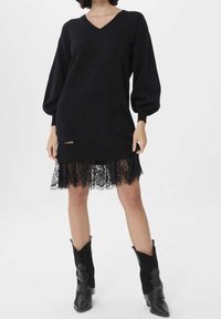 Svart stickad tröja med V-ringning, överdimensionerade ärmar och ribbad nederkant, lager-på-lager över en svart spetskjol med vågiga kanter. Svarta ankelskor.