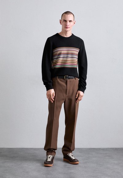 Black sweater with a horizontal multicolored stripe pattern, paired with brown tailored trousers and dark sneakers with white laces.