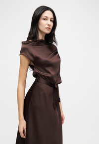 Γυναίκα με μακριά σκούρα μαλλιά που φοράει ένα αμάνικο, μεταξένιο σκούρο καφέ φόρεμα με ταιριαστή ζώνη, στέκεται μπροστά σε ένα απλό φωτεινό φόντο.