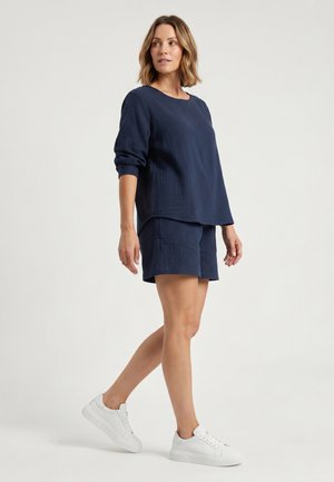 Kvinde med kort lyst brunt hår iført marineblå langærmet top og shorts, gående i hvide sneakers mod en ensfarvet lys baggrund.